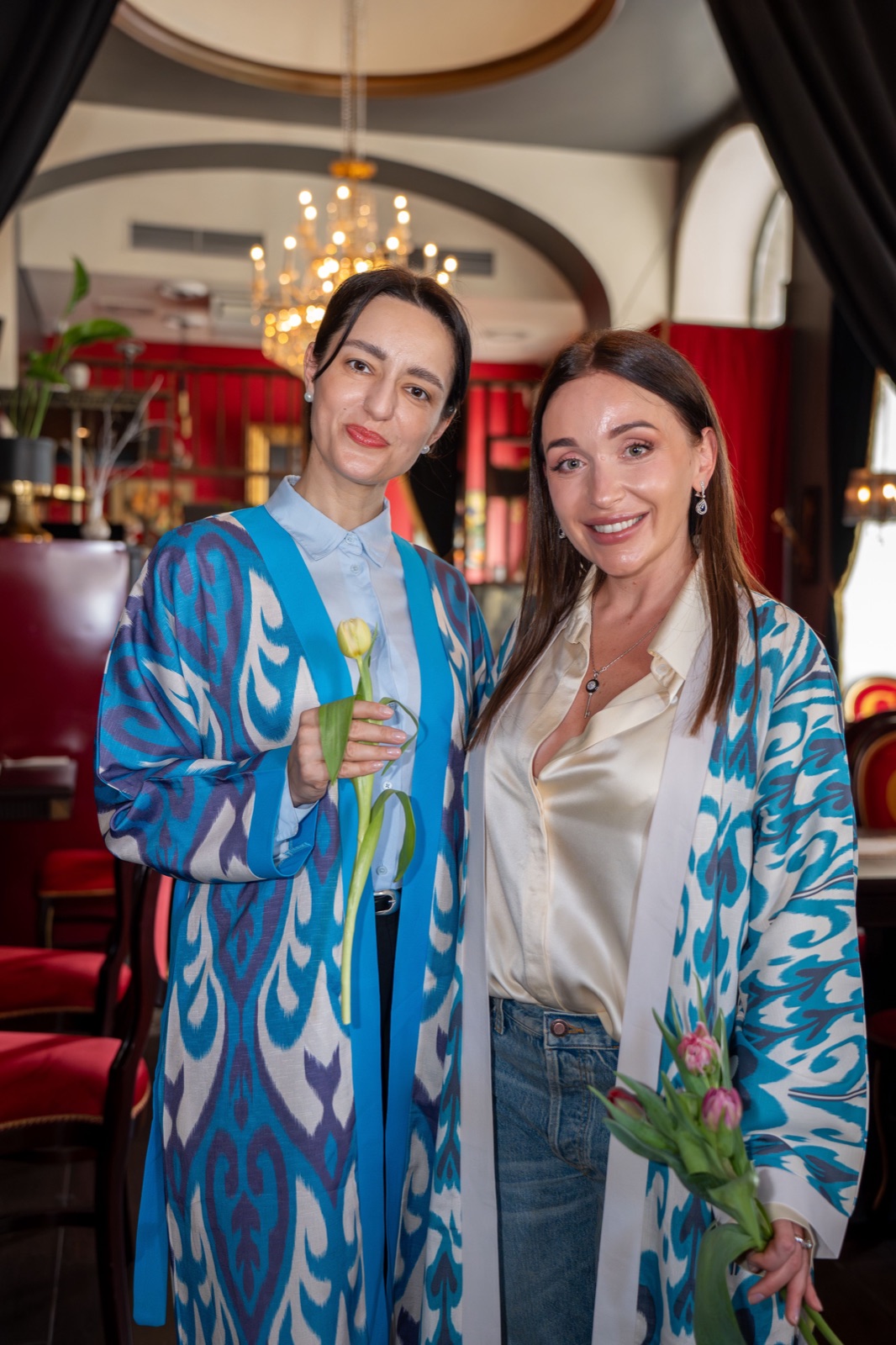 Two women in turquoise ikat coats holding tulips at a Vienna fashion event in a restaurant with chandelier — event photography by Best Alex Shots